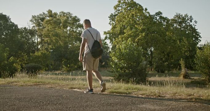 A young man with a drawstring backpack walks through a park on a sunny summer day. He walks along a paved path by grass and trees.