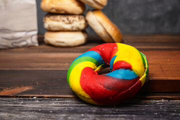 Closeup view of a rainbow bagel bread and a heap of a collection of bagel breads in various kinds with a brown paper bag on a wooden board