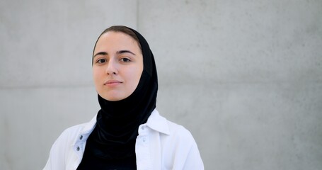 Professional businesswoman wearing hijab, standing confidently near concrete wall, expressing range of emotions while looking directly toward camera