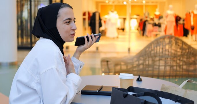 Young muslim woman wearing traditional hijab sitting at table in a shopping mall food court, talking on mobile phone and drinking coffee, enjoying a break from shopping. Free from prejudice of Muslim