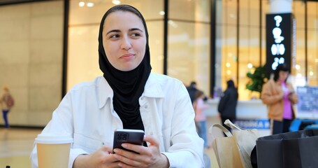 Young muslim woman wearing hijab sitting at table in shopping mall, comfortably browsing smartphone with relaxed smile, embodying modern digital lifestyle and urban connectivity
