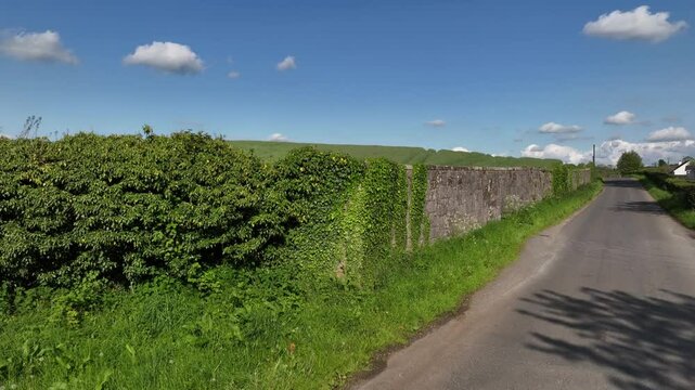 Caledon Border Wall, County Tyrone, Northern Ireland, May 2023. Drone slowly ascending over border wall, scenic road, lush farmlands and peaceful Irish countryside in summer.