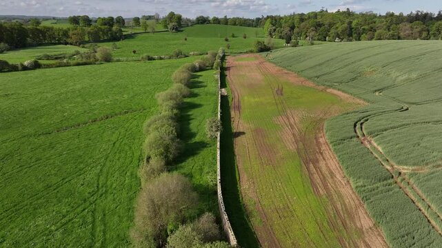 Caledon Border Wall, County Tyrone, Northern Ireland, May 2023. Drone moving forward over historic divide wall, green farmlands, vivid crop fields and rolling hills under bright sky.