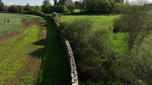 Caledon Border Wall, County Tyrone, Northern Ireland, May 2023. Drone pushes forward over stone heritage, green fields, trees, rural farmland and lush spring countryside.