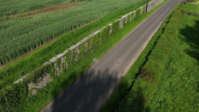 Caledon Border Wall, County Tyrone, Northern Ireland, May 2023. Drone pullback slowly over historic divide wall beside rural road and fields, aerial view of historic countryside divides and farmland.