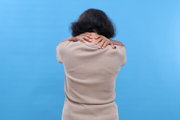 Rear view portrait of old woman touching shoulder and having shoulder or neck pain, isolated on blue background.