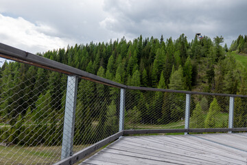 Wooden platform and railing of Tatras Tower (Dream Tower) at Štrbské Pleso, Slovakia, with forest view and ski jump structure in the distance under cloudy spring sky.
