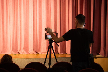 A man filming a theatre performance on a tripod while standing on a phone.