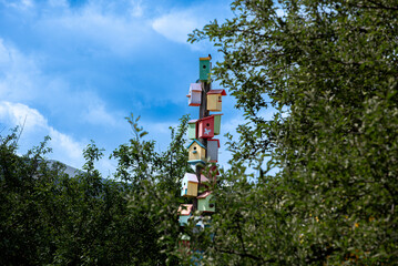 The photo shows a tall wooden pole with many colorful birdhouses stacked on top of each other. Surrounding the pole are green trees, grass, and a well with a roof in the background.