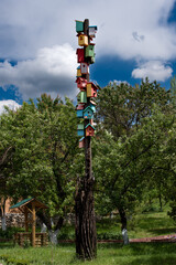 The photo shows a tall wooden pole with many colorful birdhouses stacked on top of each other. Surrounding the pole are green trees, grass, and a well with a roof in the background.