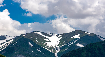 The photo shows high mountains with green slopes and melting snow. White clouds float across the blue sky, creating a contrast with the rugged and scenic mountain landscape.
