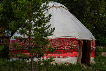 The photo shows a traditional yurt decorated with red ornamental patterns. The yurt is set among greenery, with a tree in the foreground partially covering the entrance.