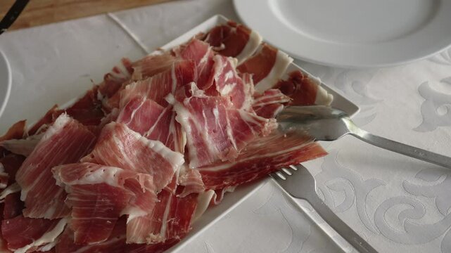 Close up of thinly sliced cured ham on a white plate with forks on a table set for a meal
