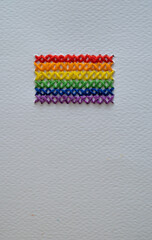 Bandera del orgullo LGBTQ+ bordada a mano con hilos de colores sobre fondo de papel blanco.
