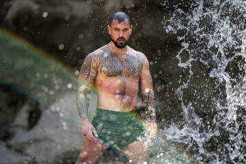 Fototapeta premium Powerful man on nature. Sexy shirtless man splashing under waterfall. Fit and strong male body in nature. Sexy model enjoying summer. Male beauty. Muscular torso with water drops. Topless guy in