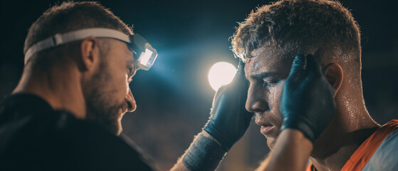 A medic with headlamp tending to a sweaty athlete with blue gloves in a dark outdoor setting after game