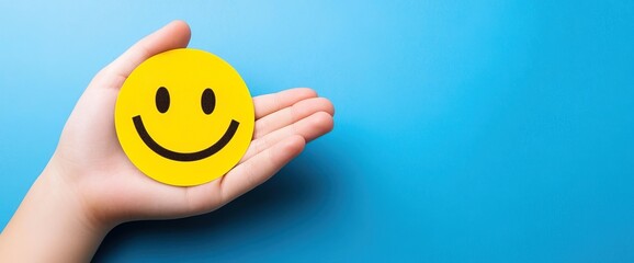 Close-up of hands holding a smiley face sticker on a blue background, with copy space for a concept of positive thinking and mental health care, or an emotional support service banner design.