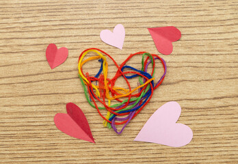 corazón de hilos con colores del arcoíris sobre madera y corazones de adorno de papel en toma cenital