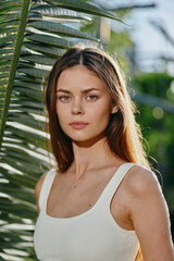 Young woman with long brown hair wearing white tank top standing near palm leaves in soft natural...