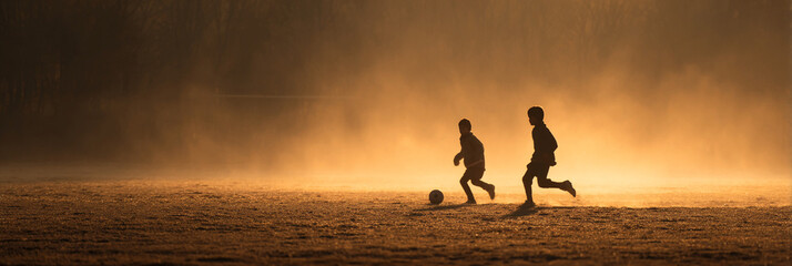 Silhouetted children playing soccer on a hazy field at sunset with golden light and dramatic sky view