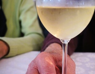 Hand von altem Mann umfasst Glas mit Weißwein auf Tisch mit weißer Stofftischdecke in Restaurant 
