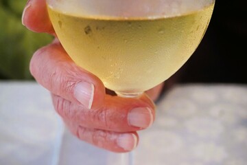Hand von altem Mann umfasst Glas mit Weißwein gefüllt auf weißer Stofftischdecke in Restaurant 