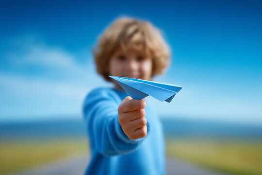 Child's hand launches blue paper plane in vivid landscape with shallow depth of field. Child is blurred behind plane in clear sky setting. Concept of creativity, adventure, childhood play