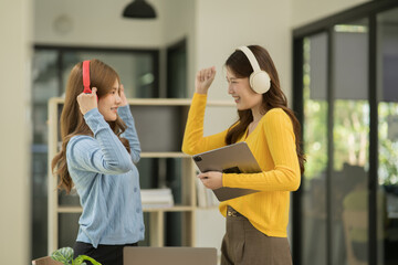 Asian woman wearing headphones standing and dancing celebrating, showing her appreciation, joy, office room