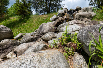 Small decorative rock waterfall with stream surrounded by grass and trees in a natural park setting.