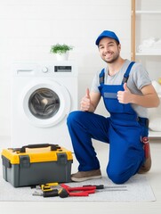 Smiling male technician in blue overalls kneeling by washing machine with toolbox and tools, giving thumbs up in bright clean laundry room