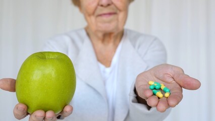 Elderly woman showing a green apple in one hand and medicine capsules in the other, highlighting the choice between natural remedies and pharmaceuticals for healthcare and wellbeing