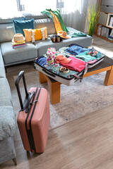 Open suitcase with summer clothes and accessories placed on coffee table and sofa in living room, symbolizing upcoming vacation and travel preparations. Copy space available at the image bottom.