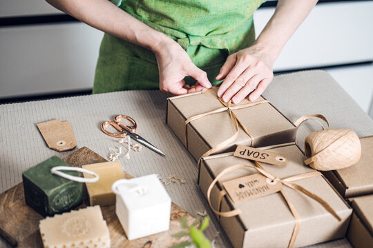 Person ties ribbon on kraft soap boxes beside artisanal bars and eco materials. Zero waste packaging, biodegradable wrap, sustainable e-commerce, green logistics