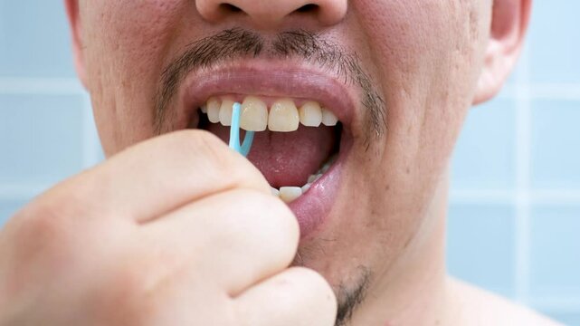 Young man brushing teeth with disposable dental floss close-up against the background of blue tiles in the bathroom.