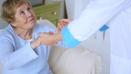 Young doctor in a white uniform holding the hands of an elderly woman seated on a sofa, providing support and compassion during her rehabilitation process in a nursing home