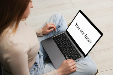 Cyber bullying. Upset teenage girl looking at laptop with text You Loser on floor indoors, closeup