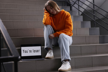 Cyber bullying. Upset teenage girl and laptop with text You Loser on stairs indoors