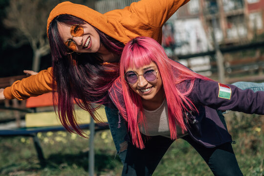  woman giving piggyback ride to girlfriend while having fun together in sunny summer day on street