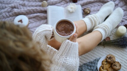 Woman in a cozy sweater and socks sipping hot chocolate while reading a book on a soft knitted blanket, surrounded by cookies, candles, and lavender
