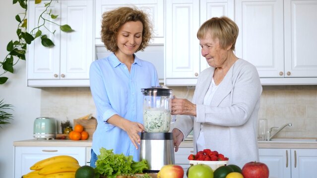 Adult daughter and her elderly mother are smiling and preparing a healthy green smoothie with a blender in a modern kitchen, surrounded by fresh fruits and vegetables
