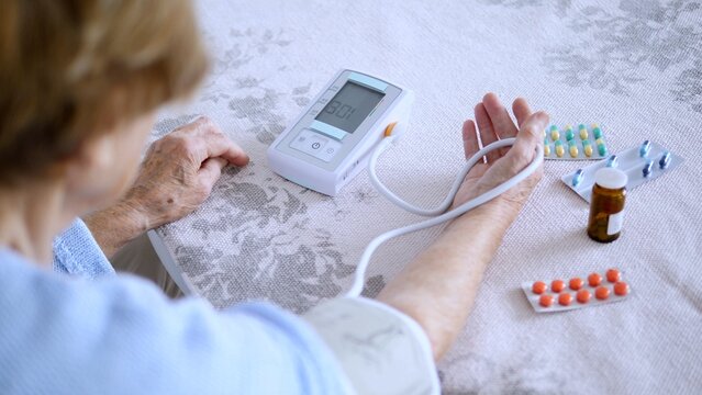 Senior woman using a blood pressure monitor at home, with various medications nearby, indicating potential health concerns and self monitoring of health conditions