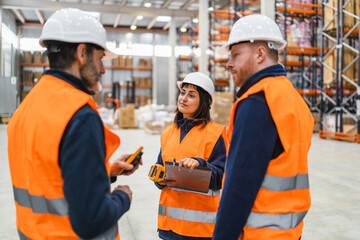 Warehouse workers wearing safety vests and hardhats are discussing logistics, holding barcode...