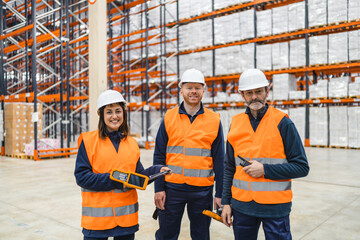 Warehouse workers wearing safety vests and hard hats are smiling and holding inventory management devices in a large warehouse