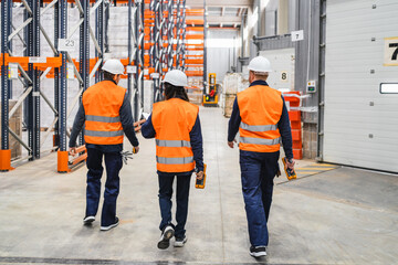 Warehouse workers wearing safety vests and helmets are walking through a large distribution center, holding handheld devices