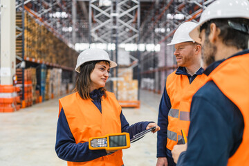 Warehouse workers wearing safety vests and helmets are discussing logistics and using handheld devices in a large warehouse