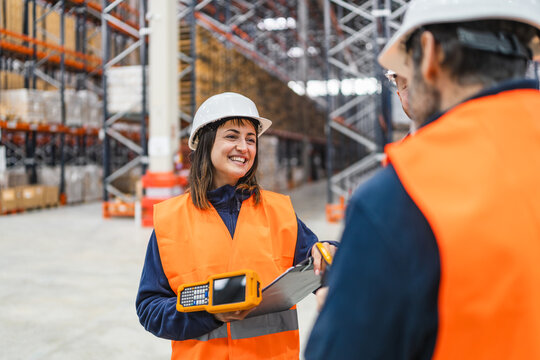 Smiling female warehouse worker holding handheld computer and clipboard discussing logistics with colleagues in large distribution center