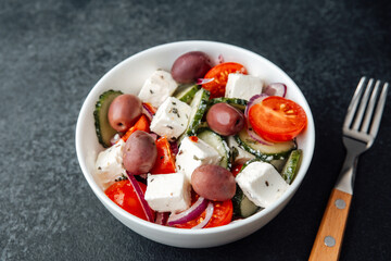 Greek salad with feta cheese in white bowl with fork on dark background