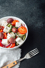 Greek salad with feta cheese olives cucumbers tomatoes onions in white bowl with fork on dark background