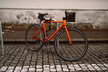 Fototapeta premium Bright orange bicycle is parked and secured to a metal bike rack on a cobblestone street. Behind it stands a historic beige building with white-framed windows, evoking a calm, urban European setting