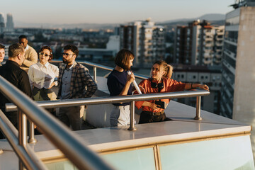A diverse group of business coworkers stands on a rooftop balcony, engaged in lively discussions....